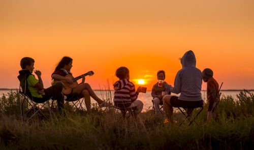 Pour fêter le solstice d’été, la famille se réunit en cercle au bord d’un lac et attend le coucher de soleil. La maman joue de la guitare, tout le monde est assis sur des chaises de camping. C’est un moment de joie et de communication avec la nature.